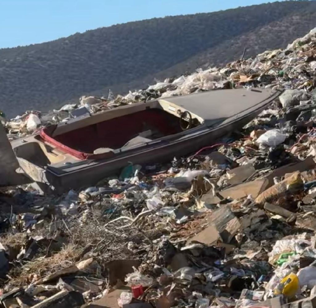 Abandoned boat discarded in a landfill, surrounded by piles of trash and debris.